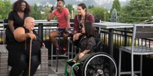 Five women sat around a table outside - one wheelchair user, one using a stick, one visually impaired and one with a proshthetic limb all of different nationalities