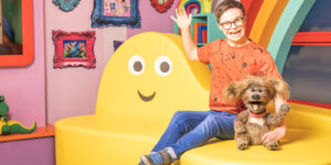 George Webster in CBeebies studio wearing orange T-shirt, blue jeans and glasses
