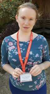 Image is a photograph of a brunette woman in a floral top, stood outdoors wearing a 'Social Distancing' ID and red lanyard