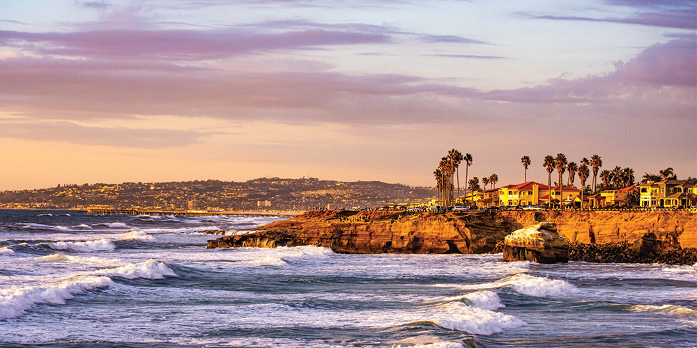 The San Diego coast seen from afar. There are cliffs with palm trees and blue sea with waves crashing. The sun is setting so there is a warm yellow glow