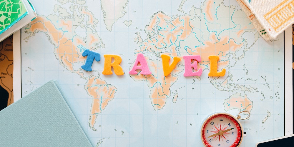 Travel image showing the words travel spelt out in yellow, blue and pink letters on a map of the world with a compass and notebooks