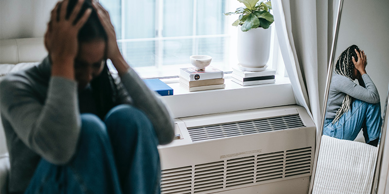 A woman sat on her bed with her head in her hands and her knees up to her chest. She is sat next to a heater below a window and window sill with books and a plant on it. She can be seen in a mirror on the other side of the room