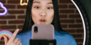 Image is a photograph of a young woman with black hair, using a selfie light and filming herself talking to the camera. Behind her is an interior brick wall with neon lights.
