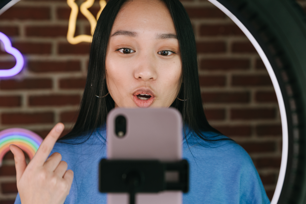 Image is a photograph of a young woman with black hair, using a selfie light and filming herself talking to the camera. Behind her is an interior brick wall with neon lights.