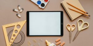 A collection of stationary and craft materials scattered across a table including a tablet, rubbers, glue, scissors, pencils, a notepad and paint palette