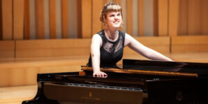 Rachel Starrit in evening attire, leaning casually on a grand piano on stage