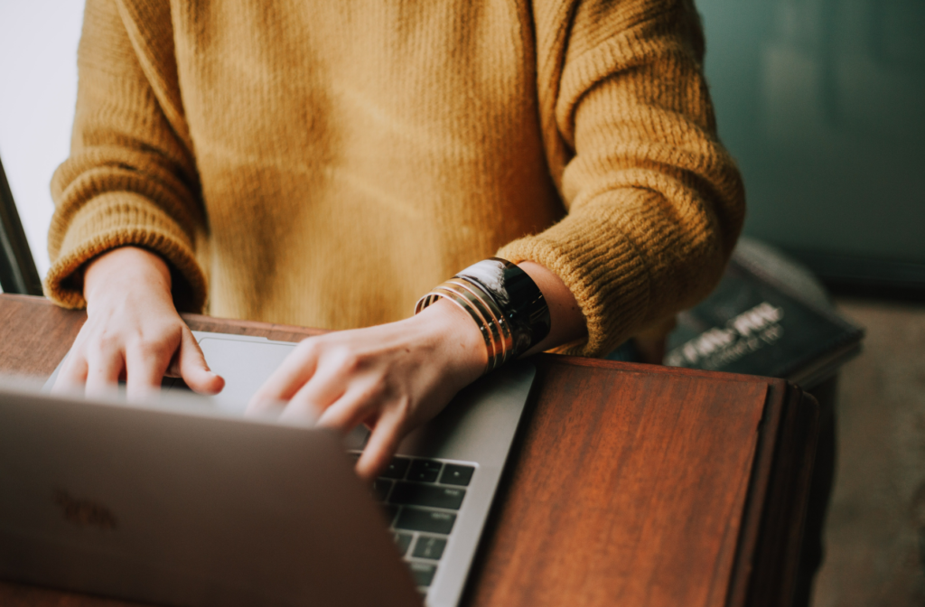 Photograph of a woman wearing a mustard-colour jumper and silver bangles, typing on a laptop on a wooden desk