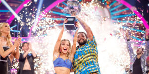 Hamza Yassin and Jowita Przystal lifting the Strictly glitterball. They are wearing brightly coloured blue and yellow outfits that they performed in for their Couple's Choice, an Afrobeats-inspired dance. There are fireworks behind them.