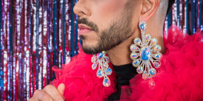 A close up of the lower part of a man's face. He has a black beard and has a pink feather boa around his neck and is wearing blue jewelled earrings