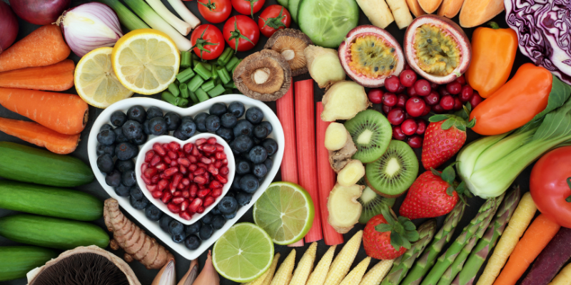 Lots of fruits and vegetables laid out with bluberries and pomegranate seeds in two white heart-shaped bowls in the middle
