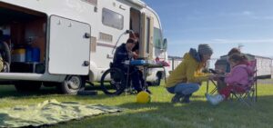 Image shows Jo crouching in front of two children in camping chairs while Rob cooks on a camping stove. They are all outside their motorhome in a field with blue sky.