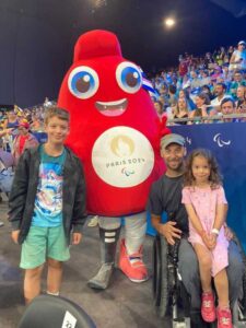 Rob and his two children meet the paralympics mascot. Xanthe sits on rob's lap and Jacob stands the other side of the mascot.