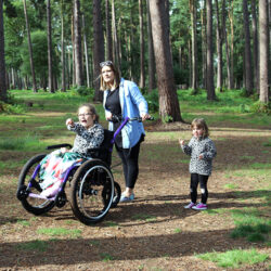 A mum with her two daughters walking in the woods. Her older daughter is in a Mountain Trike all-terrain wheelchair