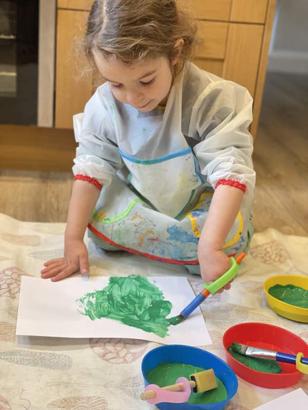 Child sits on the floor and paints using a paintbrush with eazyhold strap to support her grip.