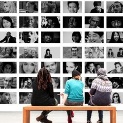 Three people sat on a bench in front of a wall of black and white portraits of people