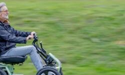 An older man with gray hair and glasses is riding a four-wheeled mobility scooter on a paved path. He is seated upright and smiling while holding the handlebars. The scooter has large tires and a supportive backrest, designed for outdoor use. The background shows a grassy field in motion blur, indicating movement.