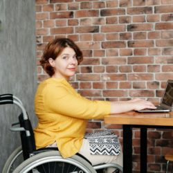 A woman in a wheelchair sits at a wooden table, working on a laptop. She is wearing a yellow top and patterned skirt, and looking toward the camera with a slight smile. The setting includes a brick wall and minimalist decor with small potted plants.