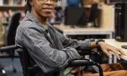 Young man in a wheelchair sitting at a computer desk in a library, smiling at the camera.
