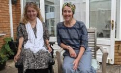 Two women sit outside a brick and glass house. One is in a power wheelchair and smiling, the other sits beside her on a wooden chair. The setting appears relaxed and friendly.