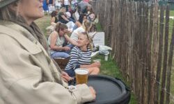 A woman in a power wheelchair watches an outdoor event from behind a fence, holding a beer on her tray. People sit on the grass beside her, facing the same direction. A crowd and scoreboard are visible in the distance under a cloudy sky.