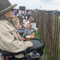 A woman in a power wheelchair watches an outdoor event from behind a fence, holding a beer on her tray. People sit on the grass beside her, facing the same direction. A crowd and scoreboard are visible in the distance under a cloudy sky.