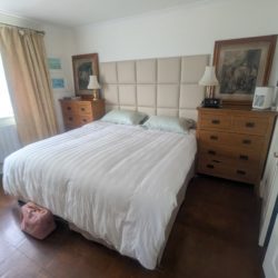 A double bed with a white duvet and green pillows, flanked by wooden bedside cabinets. A CPAP machine sits on the right cabinet next to a lamp and framed artwork.