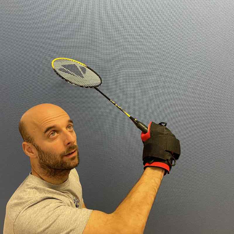 rob holds a badminton racket in a serving position, using the angled aid. He is positioned against a plain, grey background.