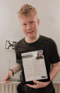 A teenage boy, Oliver, looks at the camera, holding a trophy and certificate.