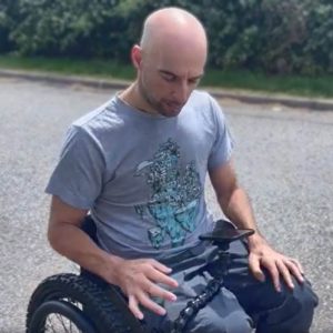 Rob is in his wheelchair, pushing along a path. he is looking down at his phone, which is being held by the Flexi Phone Holder