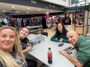 Millie, Jo, mel and rob smile at the camera for a selfie in a service station. There are drinks and snacks on the table between them.