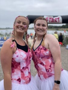 millie and jo in matching pink leotards and tutus smile at the camera. The starting line of a triathlon is in the background behind them.