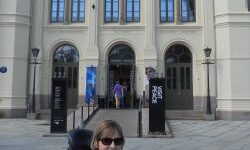 Woman in a wheelchair smiles in front of a large Oslo museum building with banners reading “Aung San Suu Kyi” and “Mother Democracy.”