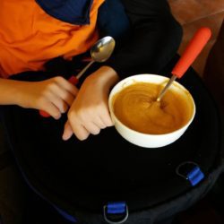 Child in an orange shirt sits with a bowl of orange soup on their lap, holding a spoon with a red handle.