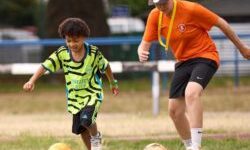 Child and football coach running drills with footballs and cones on a grass pitch during a community training session.