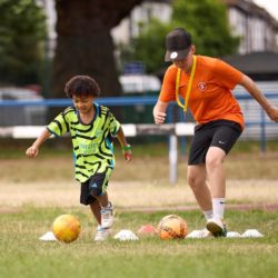 Child and football coach running drills with footballs and cones on a grass pitch during a community training session.