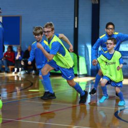 Children in blue soccer uniforms and green training bibs run after a bright green ball during an indoor soccer game while a coach watches from the side.