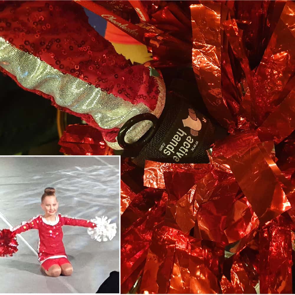 Image shows child in cheerleading uniform holding two pom poms out wide. A close up of her hand shows she is using the Limb Difference gripping aid from Active Hands to hold her pom pom.