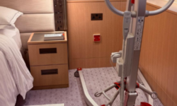 Mobile patient hoist positioned beside a hotel bed in an accessible guest room, with clear floor space for transfers.
