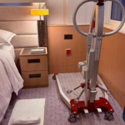 Mobile patient hoist positioned beside a hotel bed in an accessible guest room, with clear floor space for transfers.