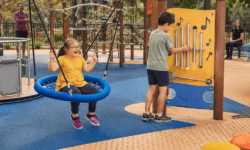 Children play in an inclusive sensory playground with a basket swing, accessible roundabout, musical play panel, rope climber, water play area and textured pathways while parents watch nearby.