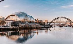 Panoramic view of the River Tyne with the Tyne Bridge and The Glasshouse reflected in calm water beneath a blue sky.