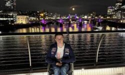Liam Virgo in his wheelchair on a footbridge over the Thames at night, with Tower Bridge, the Shard and a full moon behind him.