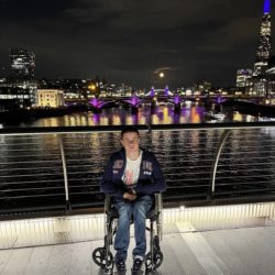 Liam Virgo in his wheelchair on a footbridge over the Thames at night, with Tower Bridge, the Shard and a full moon behind him.