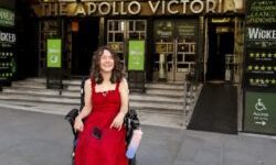Wheelchair user in a red dress outside the Apollo Victoria Theatre, with Wicked signage and an accessible ramp.