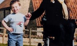 A smiling woman in a wheelchair holds hands with a young boy on a sunny lawn in front of rustic farm buildings.